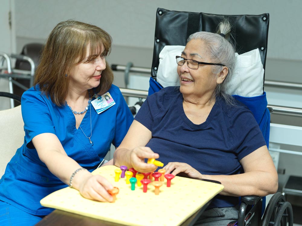 Nurse and elderly woman lying on the bed playing board games to recover from neurological issues