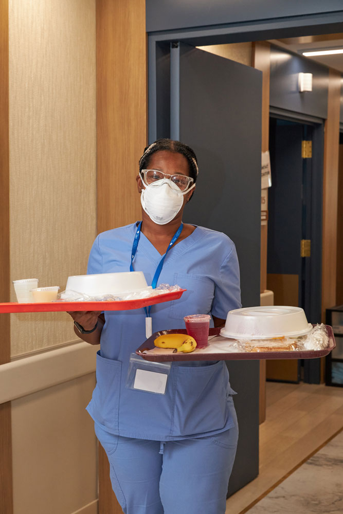 Staff holding two trays of dishes to serve food to the patient that do not raise blood pressure and is a good dysphagia diet.