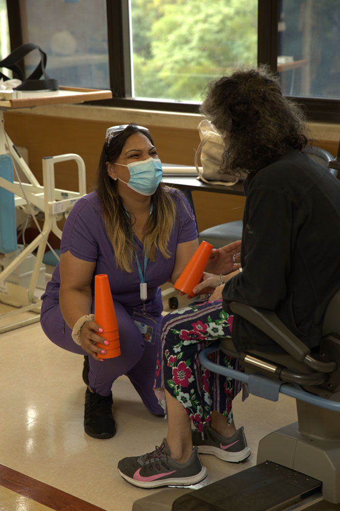 Female therapist helping senior woman sitting on a wheel chair in physical therapy for knee pain after surgery.