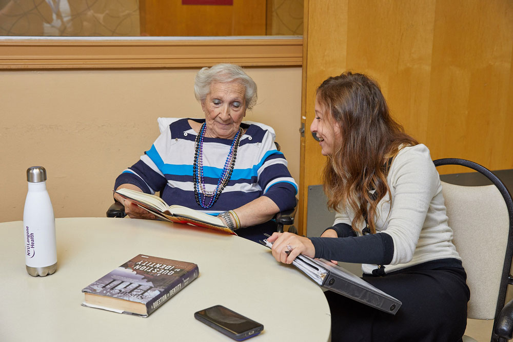 Senior woman sitting with female nurse and reading books to promote positive thinking for healing.