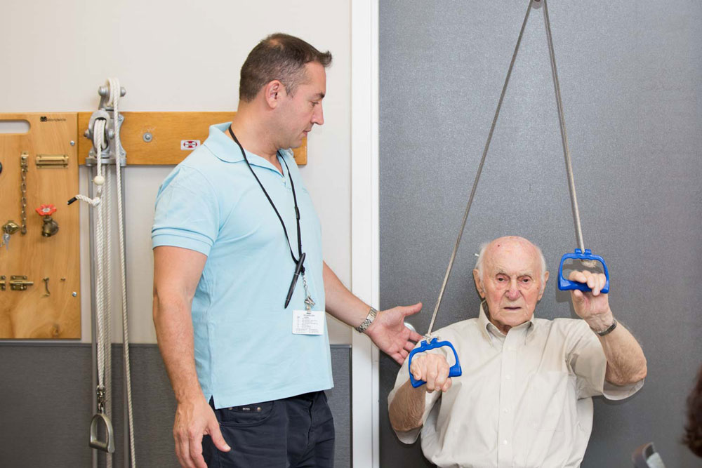 Therapist helping elderly man with exercise suffering from persistent neck and shoulder pain.