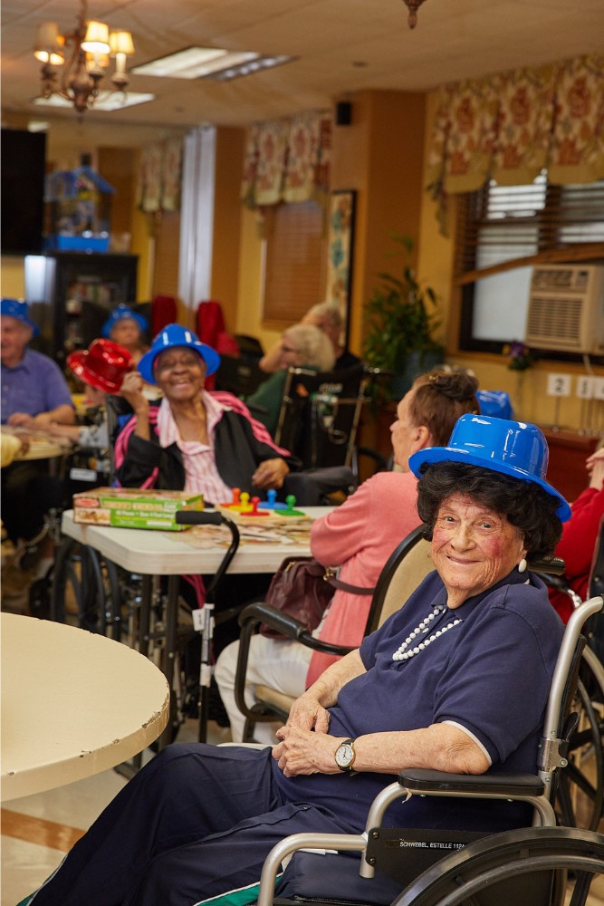 Elderly people in wheelchairs wearing colorful hats and engaging in group games at a senior center, showing the importance of wheelchair physical activities for social and mental well-being.
