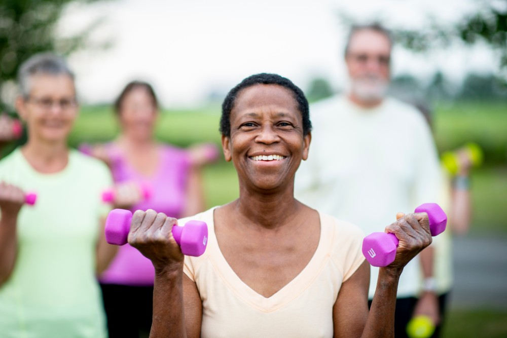 A group of senior individuals are outdoors in an open field. They are standing in a group and exercising to slow down aging process while holding dumbbells.