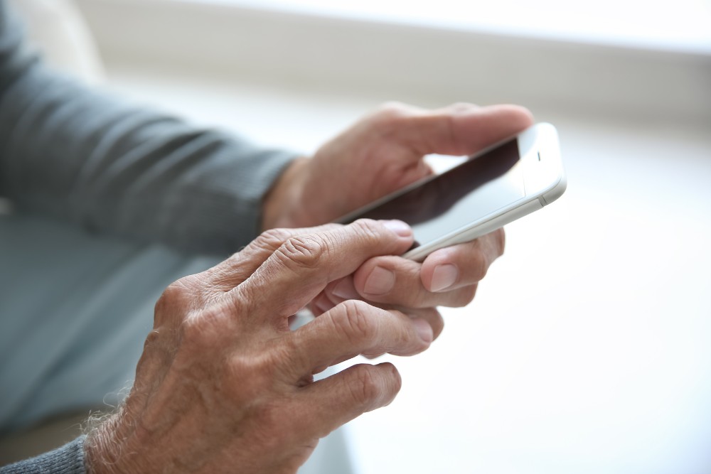 Close-up of an older adult using a smartphone with visible thumb arthritis causing discomfort while tapping the screen.