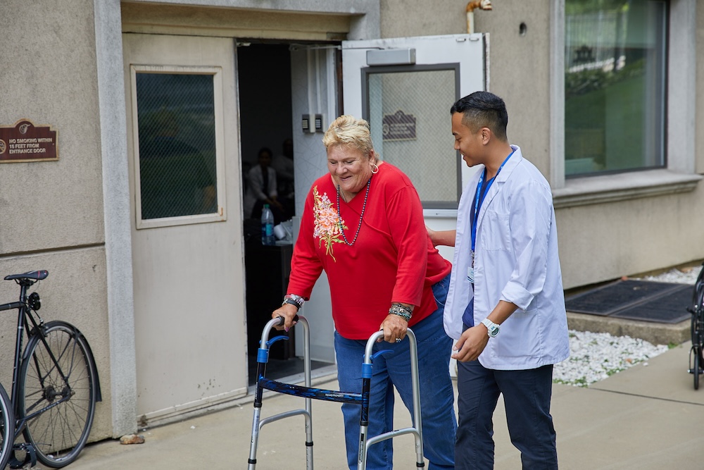 Senior woman using a walker with assistance from a healthcare professional outdoors, illustrating support for older adults experiencing balance issues during mobility rehabilitation.